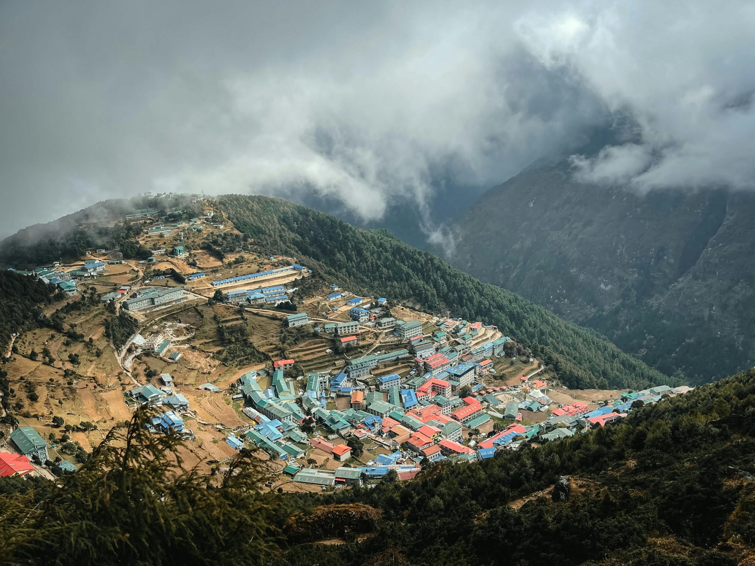 Aerial view of a traditional Sherpa village in the Everest region of Nepal, surrounded by Himalayan mountains, forests, and clouds — a popular stop on trekking routes.